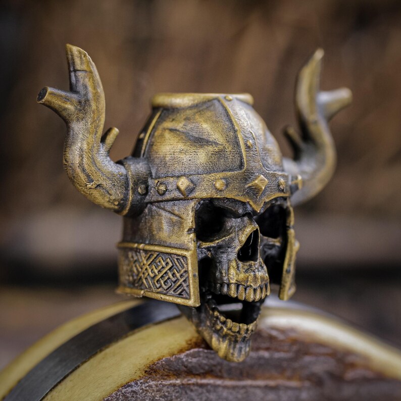 May include: A detailed, antique-style skull figurine wearing a Viking helmet with horns. The skull is black with gold accents, and the helmet is a weathered gold color. The figurine has intricate details and a textured finish.