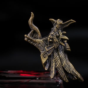 May include: A metallic bronze-toned ring featuring a detailed sculptural design of a dragon head with prominent horns, textured scales, and layered wing-like elements extending along the band, showcasing an intricate, weathered finish.