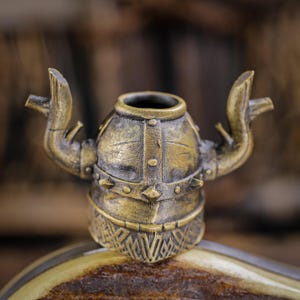 May include: A bronze-colored, Viking helmet-shaped bottle stopper. The stopper features a detailed design with a round body, a spiked collar, and two curved horns extending outwards. The helmet sits on a brown and tan surface.