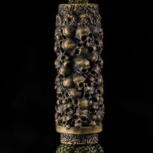 May include: A bronze-toned decorative item with a detailed design of stacked skulls. The cylindrical piece has a textured surface and is set against a black backdrop. The bottom is attached to a green, braided cord.