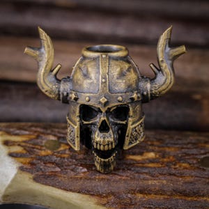 May include: A detailed, antique-style skull figurine wearing a Viking helmet with horns. The skull is bronze-colored, with intricate details on the helmet and face. The figurine is set against a blurred wooden background.