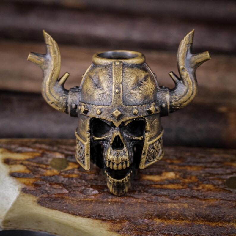 May include: A detailed, antique-style skull figurine wearing a Viking helmet with horns. The skull is bronze-colored, with intricate details on the helmet and face. The figurine is set against a blurred wooden background.
