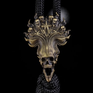 Peut inclure: Un pendentif en métal couleur laiton représentant un crâne central à la mâchoire ouverte, entouré de crânes plus petits et de motifs tourbillonnants en forme de flammes. Il est fixé à un cordon tressé noir épais qui s'étend vers le haut.