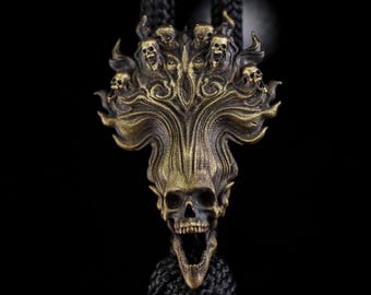 May include: A brass-toned metal pendant featuring a central skull with an open jaw, surrounded by smaller skulls and flame-like swirling patterns. The pendant is attached to a thick, black braided cord that extends upward and downward.