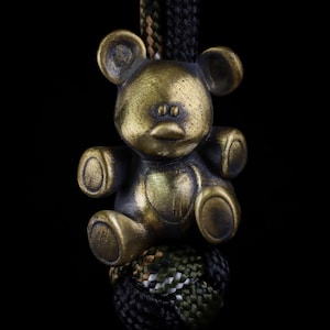 May include: A brass-toned metal bear charm with a weathered finish, featuring rounded ears, a small snout, and a heart-shaped detail on the chest. The bear is attached to a thick, braided paracord lanyard with black, green, and white patterns.