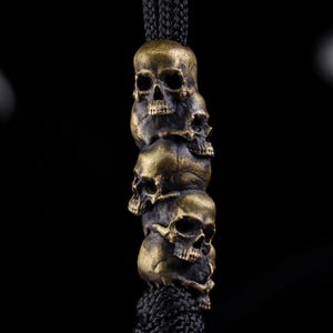 May include: A vertical stack of four metallic brass-colored skull beads threaded onto a thick black braided cord. Each skull features detailed eye sockets, nasal cavities, and teeth, with a weathered, dark patina finish across the metal surface.