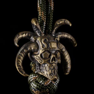 May include: A bronze-colored skull pendant with curved horns and a jester-like design. The skull has intricate details and is attached to a green, brown, and white braided cord. The background is black.