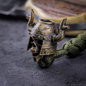 May include: A bronze-colored skull bead with a Viking helmet design. The bead features a detailed skull face, a helmet with horns, and decorative patterns. It is attached to a green braided cord, creating a unique accessory.