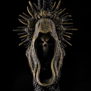 May include: A bronze-colored pendant featuring a skull within a hooded cloak, adorned with a spiky halo design. The pendant is suspended from a black cord, set against a dark background. The intricate details and gothic theme are prominent.