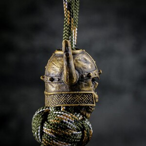 May include: A bronze-colored Viking helmet charm with a skull face design. The helmet has a horn and is attached to a green, brown, and white braided cord. The charm is detailed and has an aged appearance.