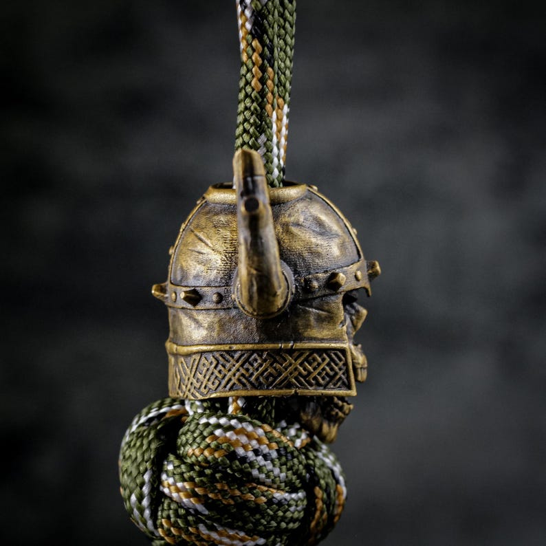 May include: A bronze-colored Viking helmet charm with a skull face design. The helmet has a horn and is attached to a green, brown, and white braided cord. The charm is detailed and has an aged appearance.