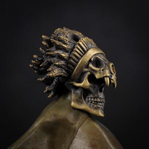 Op de afbeelding: Een bronzen schedelsculptuur met een gevederde hoofdtooi. De schedel heeft gedetailleerde tanden en oogkassen, met een band om het voorhoofd. De veren zijn ingewikkeld ontworpen en de sculptuur heeft een donkere achtergrond.