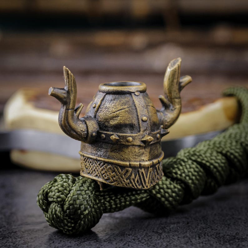 May include: A bronze-colored Viking helmet bead with raised arms, detailed with rivets and etched patterns. The bead is attached to a green braided cord, creating a unique accessory. The background is blurred, highlighting the bead's intricate design.