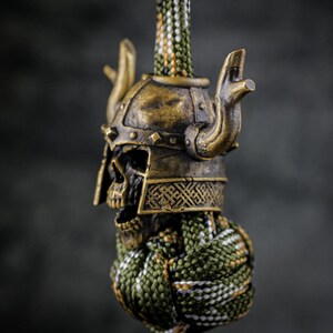 May include: A bronze-colored skull wearing a Viking helmet with horns. The skull is attached to a green, white, and brown braided cord. The helmet has intricate details and a weathered appearance, suggesting a vintage or antique aesthetic.