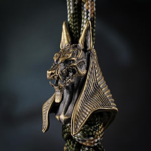 May include: A detailed bronze Anubis pendant, depicting the jackal-headed god. The pendant hangs from a green, brown, and beige braided cord. This Anubis pendant is a unique piece of jewellery, with a detailed design.