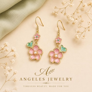 May include: Gold-tone drop earrings featuring a small pink flower with a turquoise center above a larger pink flower with a white pearl center and a green leaf accent. The text reads ANGELES JEWELRY TIMELESS BEAUTY, MADE FOR YOU.