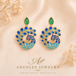 May include: A pair of gold-toned dangle earrings featuring a peacock motif with blue and turquoise enamel detailing, a small pink stone for the eye, and a teardrop-shaped green stone at the top. The text reads ANGELES JEWELRY TIMELESS BEAUTY, MADE FOR YOU.