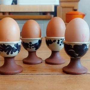 May include: Four brown eggs in ceramic egg cups. The cups have a cream-colored base with a dark blue floral design and a brown pedestal. The eggs are light brown and sit on a wooden surface.