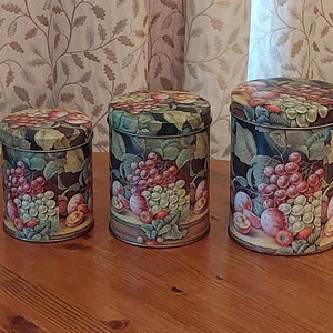 Puede incluir: Juego de tres botes metálicos vintage con tapas. Cada bote presenta un diseño pintado de uvas, ciruelas y hojas en tonos rojos, verdes y morados sobre un fondo oscuro. Los botes son cilíndricos y de diferentes tamaños.