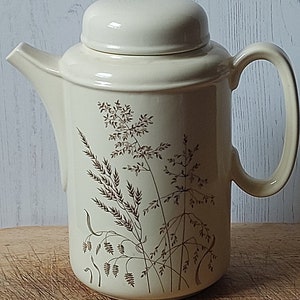 May include: Cream-coloured ceramic teapot with a lid and a curved handle. The teapot features a brown floral design. The spout is on the left side, and the handle is on the right. The teapot is on a wooden surface.
