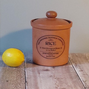 May include: A tan ceramic canister with a lid and a knob, labeled "The Original Suffolk Canister" with "Rice" printed on it. A yellow lemon sits to the left of the canister. The canister is made by Henry Watson Pottery, founded in 1600.
