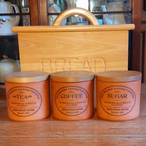 May include: A wooden bread box with a handle and the word "BREAD" carved into the front. Three orange canisters with wooden lids are labeled "TEA", "COFFEE", and "SUGAR". Each canister has a circular label.