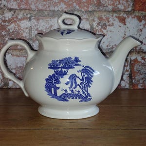 Puede incluir: Una tetera de cerámica blanca con un diseño azul de sauce. La tetera tiene un pico curvo, un asa y una tapa con un lazo. El diseño azul representa una escena de paisaje con árboles, un puente y figuras.