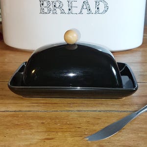 May include: A rectangular black ceramic butter dish with a matching domed lid and a small, round, light-colored wooden knob handle. The dish is accompanied by a silver-toned metal butter knife with a pointed, split tip for serving.