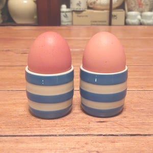 May include: Two pink eggs sit in blue and white striped ceramic egg cups on a wooden surface. The egg cups have a classic design, with alternating bands of blue and white. The eggs are a soft pink color.