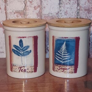 Puede incluir: Dos botes de cerámica blanquecina con tapas de madera. Uno presenta un diseño de hoja azul, el otro un helecho blanco. Ambos tienen un borde rojo y el texto "Tea" y "Sugar".