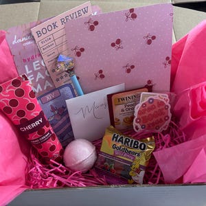 May include: A pink gift box filled with cherry-themed items. Includes a cherry-scented hand cream, a bath bomb, a pen, a book review card, a tea bag, a sticker, and a bag of gummy bears. The box is lined with pink tissue paper.