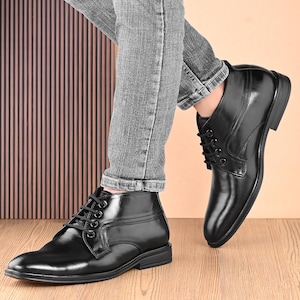 May include: Black leather ankle boots with a lace-up design. The boots have a polished finish and a low heel. The image shows the boots from a low angle, highlighting their sleek design. The boots are worn with gray jeans.