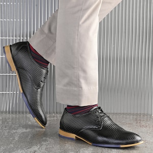 May include: A pair of black leather lace-up shoes with a woven pattern. The shoes have a light brown sole and a blue trim around the edge. The shoes are being worn with beige trousers and red and blue striped socks.