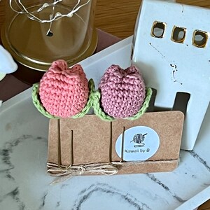 Puede incluir: Dos tulipanes rosas de crochet con hojas verdes unidos a pinzas para el pelo de metal dorado. Las pinzas para el pelo están en una tarjeta marrón con un círculo blanco que dice "Kawaii by B" y una pequeña imagen de hilo.