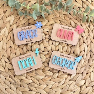 May include: Four wooden name plaques with the names Grace, Olivia, Noah, and George in colourful 3D lettering. Each plaque has the phrase "look what made" and a decorative element like a flower, bow, or rocket.