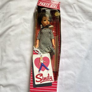 May include: A Sindy doll in original packaging labeled Party Girl. The doll has dark hair and wears a black and white striped dress with a black collar. A white tag attached to the box features a pink heart graphic with a cartoon figure and the text Sindy.