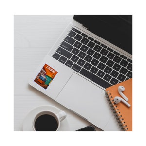 May include: A laptop computer with a sticker of a vintage tram in Lisbon, Portugal. The sticker is orange, yellow, and blue with the word "LISBON" in white text. A cup of coffee and a notebook with earbuds are also in the image.