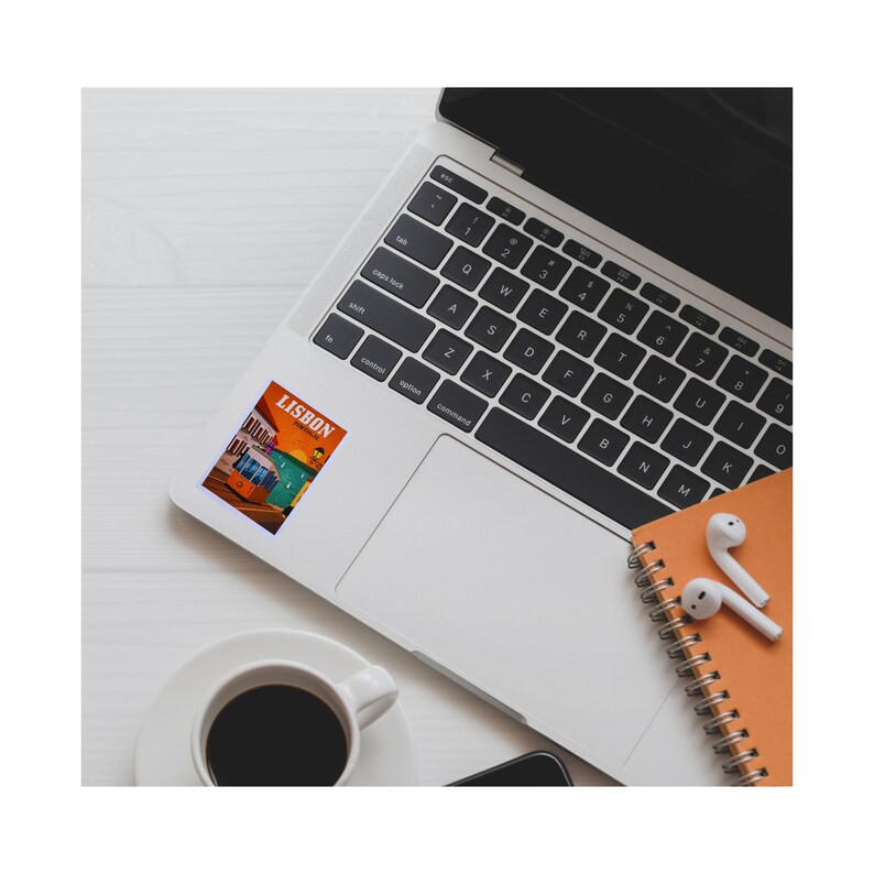 May include: A laptop computer with a sticker of a vintage tram in Lisbon, Portugal. The sticker is orange, yellow, and blue with the word "LISBON" in white text. A cup of coffee and a notebook with earbuds are also in the image.