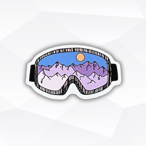 May include: A sticker of a pair of ski goggles with a mountain landscape and a sun visible through the lenses. The goggles are white with black straps and a blue, purple, and white mountain scene inside.