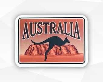 May include: A rectangular sticker featuring a vintage-style design. The word "AUSTRALIA" is arched across the top in a bold font. Below, a black kangaroo silhouette leaps over a red rock formation, set against a gradient orange background.