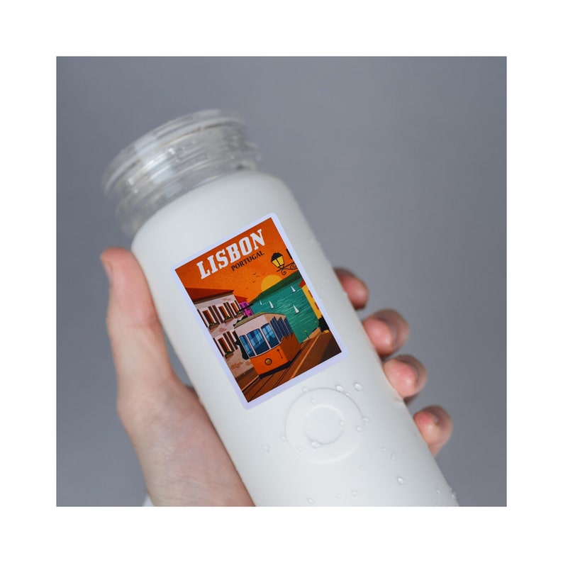 May include: White reusable water bottle with a sticker featuring a colorful illustration of a tram in Lisbon, Portugal. The sticker says "Lisbon, Portugal".