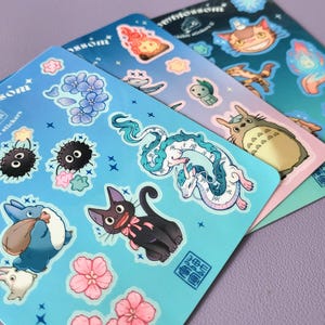 May include: A set of blue, pink, and green sticker sheets featuring characters from the animated film My Neighbor Totoro. The stickers include Totoro, the Catbus, and other characters from the film. The stickers are printed with a glossy finish and have a whimsical, cartoon style.