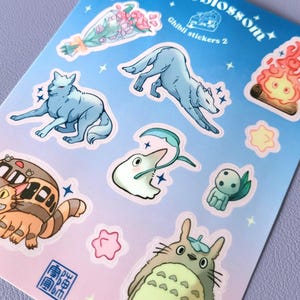 May include: A sheet of stickers featuring characters from Studio Ghibli films. The stickers are on a blue and pink gradient background with white outlines. The stickers include Totoro, No Face, the wolf from Princess Mononoke, and a cat bus.
