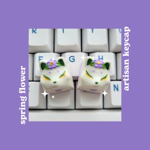 May include: Two white artisan keycaps with a fox design and purple flowers. The keycaps are on a white keyboard.