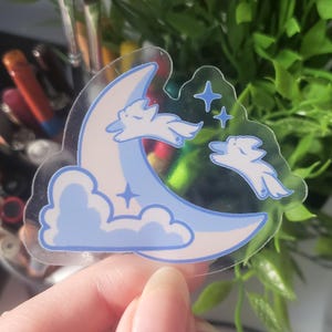 May include: A clear sticker featuring a light blue crescent moon, clouds, and stars. Two white cartoon animals are leaping across the moon. The sticker has a holographic effect.