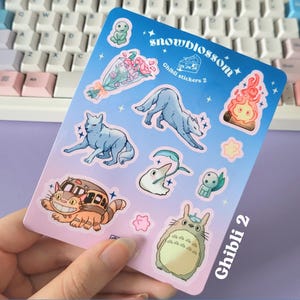 May include: A sheet of stickers featuring characters from Studio Ghibli films. The stickers are on a blue and pink background with white stars. The text "snowblossom Ghibli stickers 2" is at the top of the sheet. The stickers include Totoro, No Face, the wolf from Princess Mononoke, and other characters.