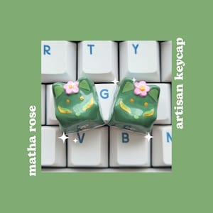 Peut inclure: Deux touches artisanales vertes et dorées avec une fleur rose et un visage blanc sur un clavier. Les touches sont étiquetées "G".