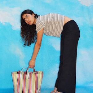 Könnte beinhalten: Eine Person trägt ein gestreiftes Shirt und eine schwarze Hose und hält eine große, farbenfrohe, gestreifte Tragetasche. Die Tasche hat eine gewebte Textur und ist aus Stoff.