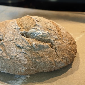 May include: A freshly baked loaf of sourdough bread with a rustic crust and a slightly cracked surface. The bread is sitting on a piece of parchment paper.