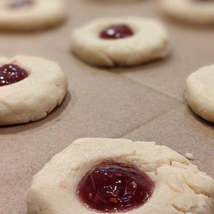May include: Close-up of a batch of homemade cookies with a red raspberry jam filling. The cookies are on a brown parchment paper-lined baking sheet.